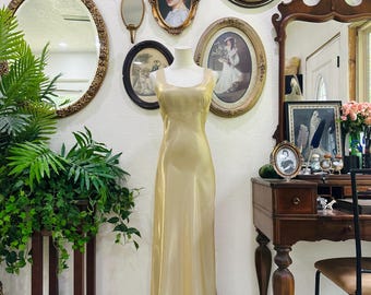 May include: A floor-length, champagne-colored gown on a mannequin. The dress has a simple, elegant design with a slight sheen. The background features antique mirrors and framed portraits, creating a vintage aesthetic.