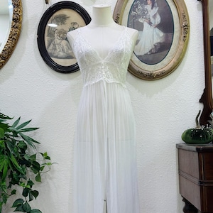 May include: A floor-length white nightgown featuring a sheer, flowing skirt and a bodice detailed with intricate white lace. The garment has a deep V-neckline and thin shoulder straps, displayed on a mannequin to show the full length and drape.