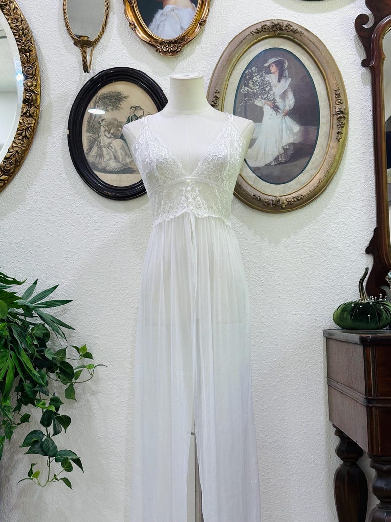 May include: A floor-length white nightgown featuring a sheer, flowing skirt and a bodice detailed with intricate white lace. The garment has a deep V-neckline and thin shoulder straps, displayed on a mannequin to show the full length and drape.