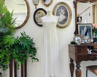 May include: A white, vintage-style dress on a mannequin, displayed against a wall adorned with antique mirrors and framed portraits. The dress has a delicate, lacy bodice and a flowing skirt. A dark wooden plant stand with greenery is in the foreground.