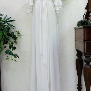 May include: A floor-length white robe featuring a wrap-style front with a matching tie belt at the waist. The garment has long, flowing bell sleeves and a V-neckline, crafted from a sheer, lightweight fabric that drapes down to the floor.