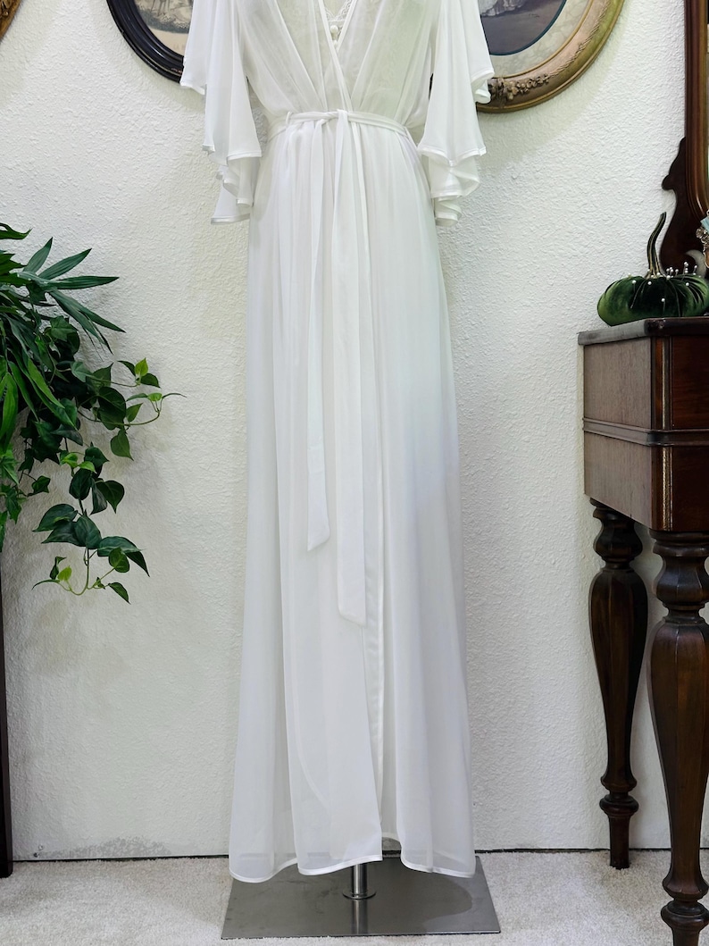 May include: A floor-length white robe featuring a wrap-style front with a matching tie belt at the waist. The garment has long, flowing bell sleeves and a V-neckline, crafted from a sheer, lightweight fabric that drapes down to the floor.