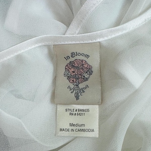 May include: A white sheer garment featuring a fabric clothing tag. The tag displays the text In Bloom by jonquil, a floral illustration, STYLE # SWB035, RN # 64211, Medium, and MADE IN CAMBODIA. The fabric has a smooth, semi-transparent texture.