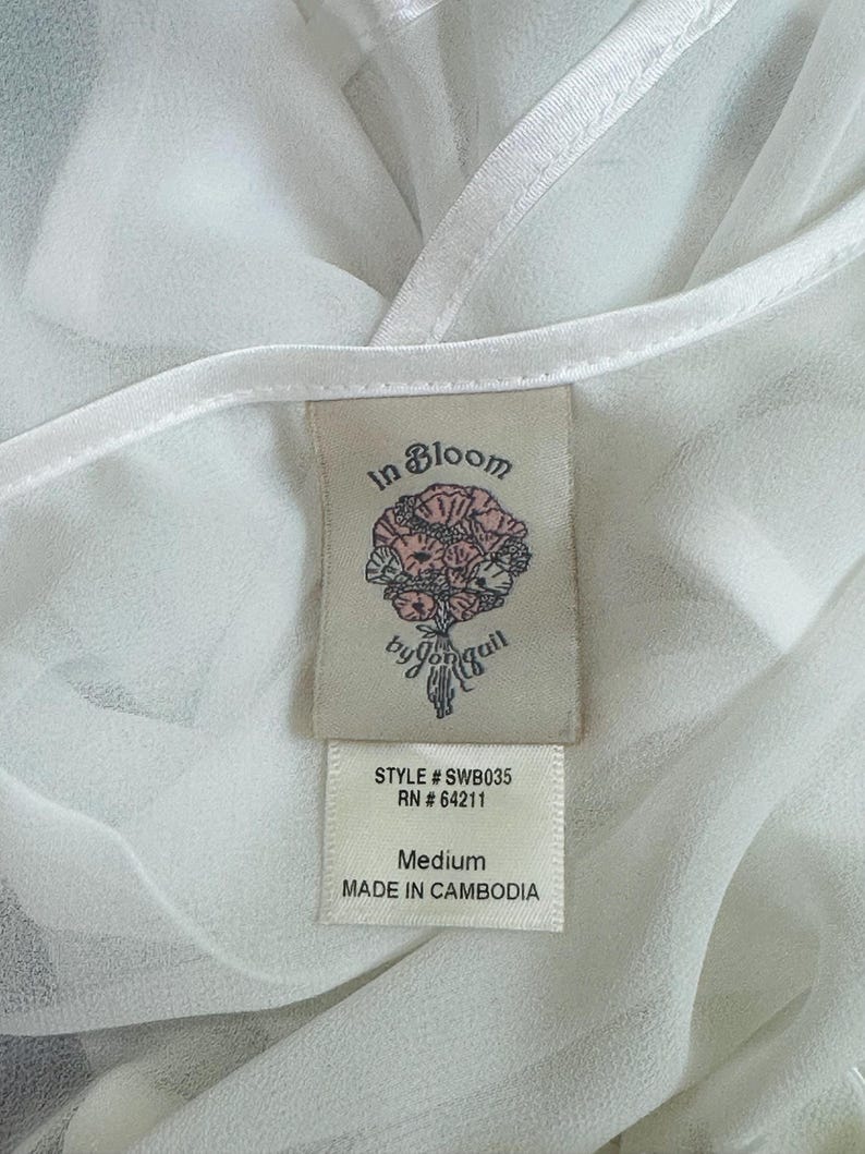 May include: A white sheer garment featuring a fabric clothing tag. The tag displays the text In Bloom by jonquil, a floral illustration, STYLE # SWB035, RN # 64211, Medium, and MADE IN CAMBODIA. The fabric has a smooth, semi-transparent texture.