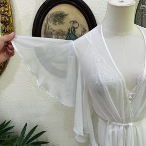 May include: A sheer white robe featuring wide, flowing bell sleeves and a delicate lace trim along the front opening. The garment is tied at the waist with a matching fabric sash and is displayed on a mannequin, showcasing the lightweight fabric.