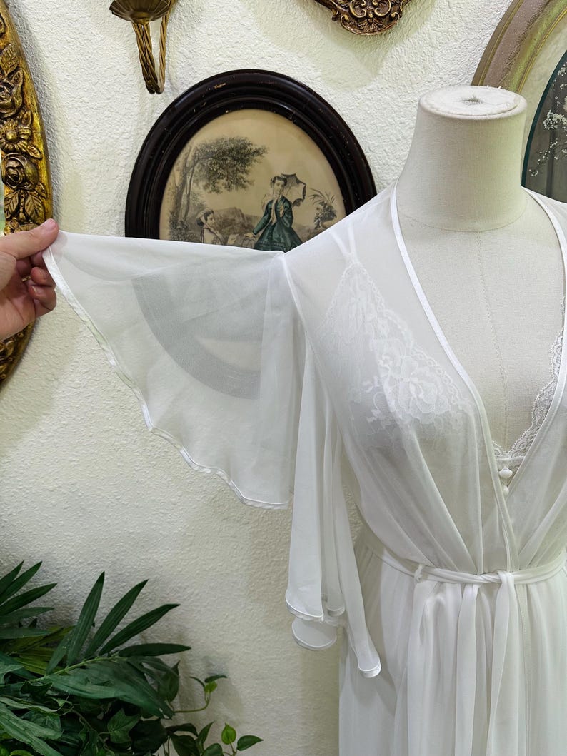 May include: A sheer white robe featuring wide, flowing bell sleeves and a delicate lace trim along the front opening. The garment is tied at the waist with a matching fabric sash and is displayed on a mannequin, showcasing the lightweight fabric.