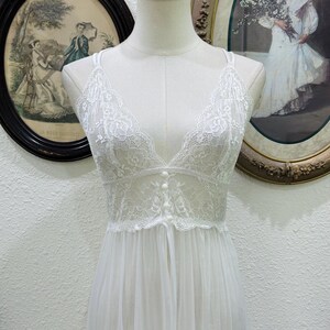 May include: A white sleeveless nightgown featuring a sheer lace bodice with a deep V-neckline and three small decorative buttons down the center. The garment has thin shoulder straps and a flowing, semi-sheer skirt made of lightweight fabric.