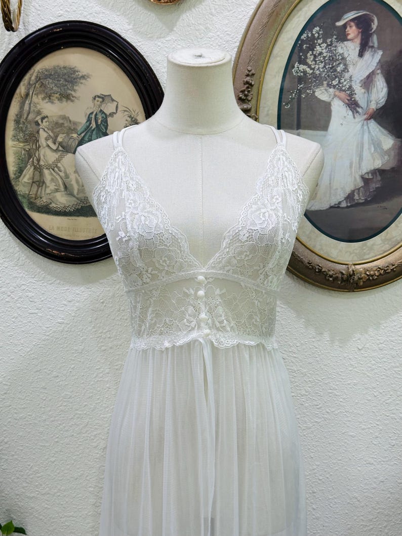 May include: A white sleeveless nightgown featuring a sheer lace bodice with a deep V-neckline and three small decorative buttons down the center. The garment has thin shoulder straps and a flowing, semi-sheer skirt made of lightweight fabric.