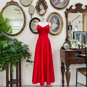 May include: A vibrant red, floor-length dress with spaghetti straps is displayed on a mannequin. The dress has a textured pattern and a V-neckline. The background features antique mirrors and framed artwork, creating a vintage aesthetic.