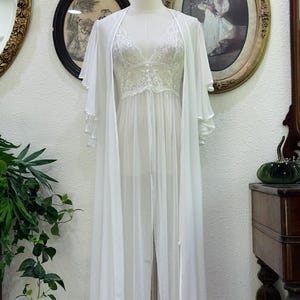 May include: A white floor-length sheer robe with wide flutter sleeves paired with a matching white lace camisole featuring a V-neckline and delicate floral embroidery. The ensemble is displayed on a mannequin, showcasing the layered fabric.