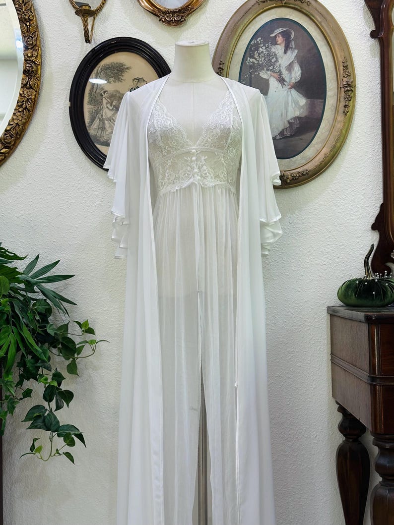 May include: A white floor-length sheer robe with wide flutter sleeves paired with a matching white lace camisole featuring a V-neckline and delicate floral embroidery. The ensemble is displayed on a mannequin, showcasing the layered fabric.