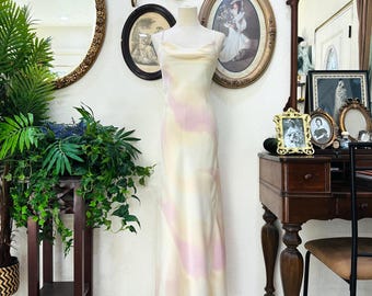 May include: A floor-length slip dress featuring a cowl neckline and thin spaghetti straps. The fabric is a light cream color with soft, abstract watercolor-style swirls in shades of pale pink and yellow throughout the garment.