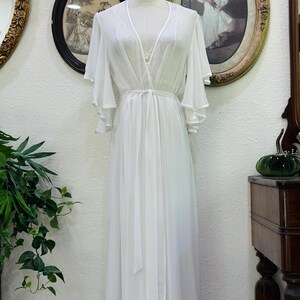 May include: A floor-length white robe featuring sheer fabric, wide flutter sleeves, and a wrap-style front with a matching tie belt. The garment includes delicate lace detailing along the V-neckline and is displayed on a mannequin.
