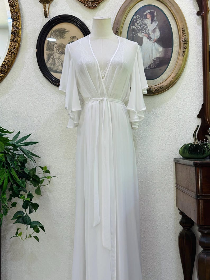 May include: A floor-length white robe featuring sheer fabric, wide flutter sleeves, and a wrap-style front with a matching tie belt. The garment includes delicate lace detailing along the V-neckline and is displayed on a mannequin.