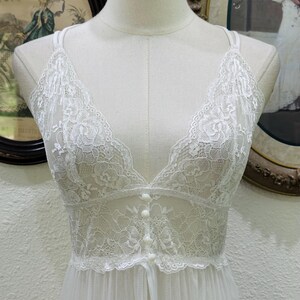 May include: A white sleeveless nightgown featuring a deep V-neckline and a bodice crafted from intricate floral lace. The front of the bodice is adorned with a vertical row of four small, round, matching white buttons, transitioning into a sheer, flowing skirt.