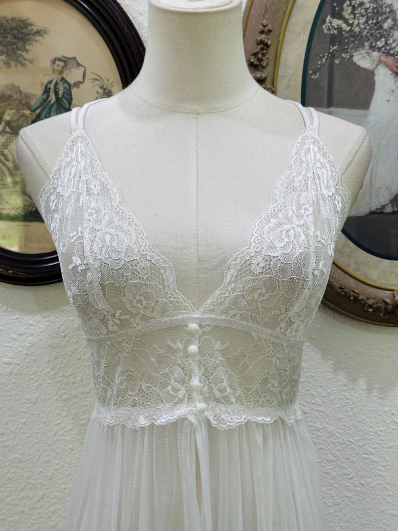 May include: A white sleeveless nightgown featuring a deep V-neckline and a bodice crafted from intricate floral lace. The front of the bodice is adorned with a vertical row of four small, round, matching white buttons, transitioning into a sheer, flowing skirt.