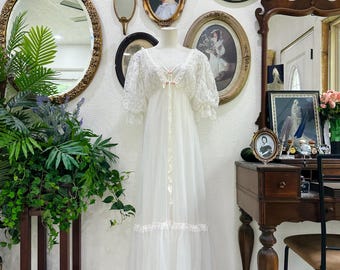 May include: A vintage white lace gown with short sleeves and a flowing skirt. The dress features delicate lace detailing on the bodice and sleeves, with a ribbon accent at the waist. The gown is displayed on a mannequin in front of a wall with antique mirrors and framed portraits.