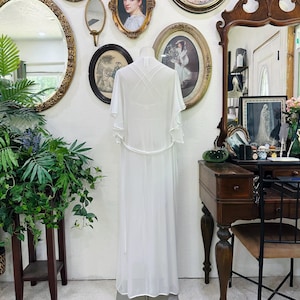 May include: A floor-length white sheer robe featuring short flutter sleeves, a thin waist tie, and a crisscross strap design on the back. The garment is displayed on a mannequin, showcasing a lightweight, flowing silhouette with a solid white base.