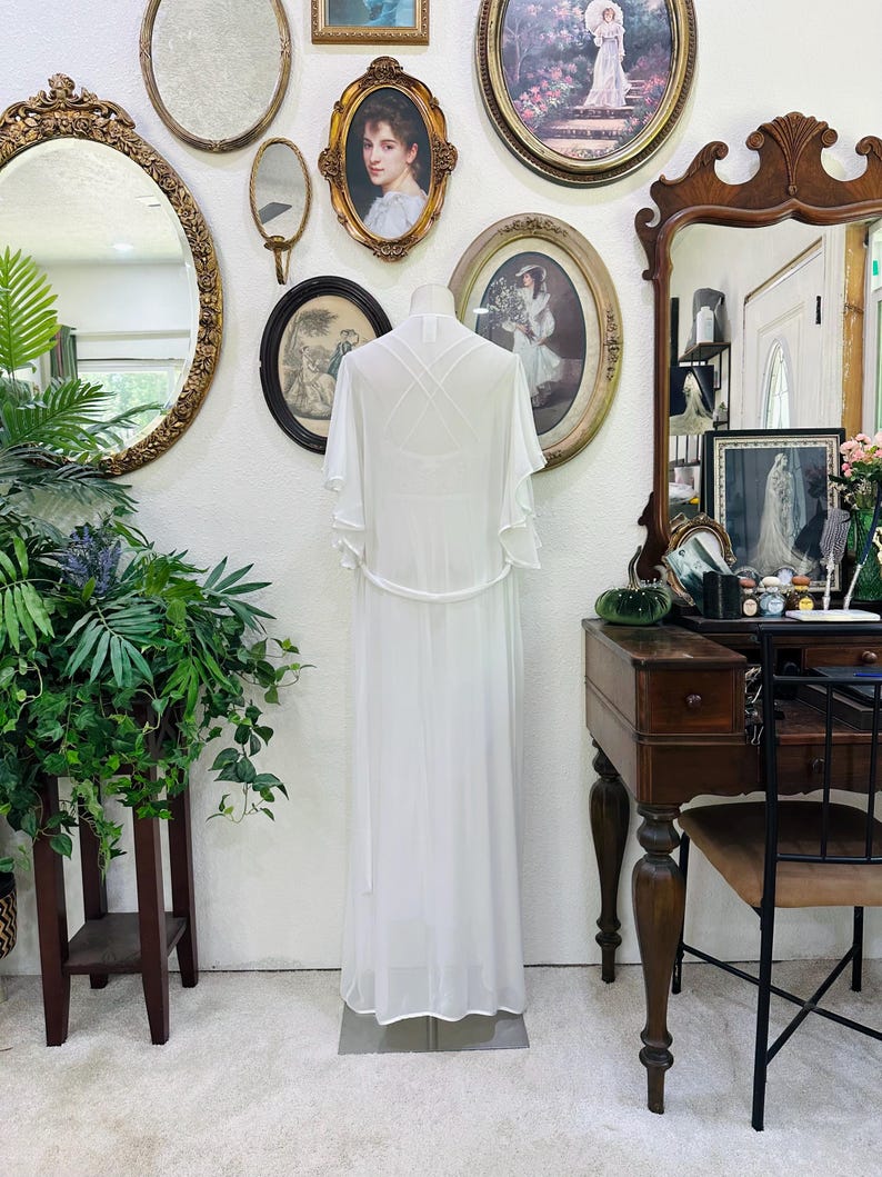 May include: A floor-length white sheer robe featuring short flutter sleeves, a thin waist tie, and a crisscross strap design on the back. The garment is displayed on a mannequin, showcasing a lightweight, flowing silhouette with a solid white base.