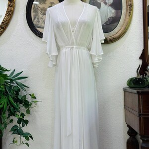 May include: A floor-length white robe featuring sheer fabric, a deep V-neckline, and wide, flowing bell sleeves. The garment includes a matching tie belt at the waist and delicate lace detailing along the neckline and shoulders, displayed on a mannequin.