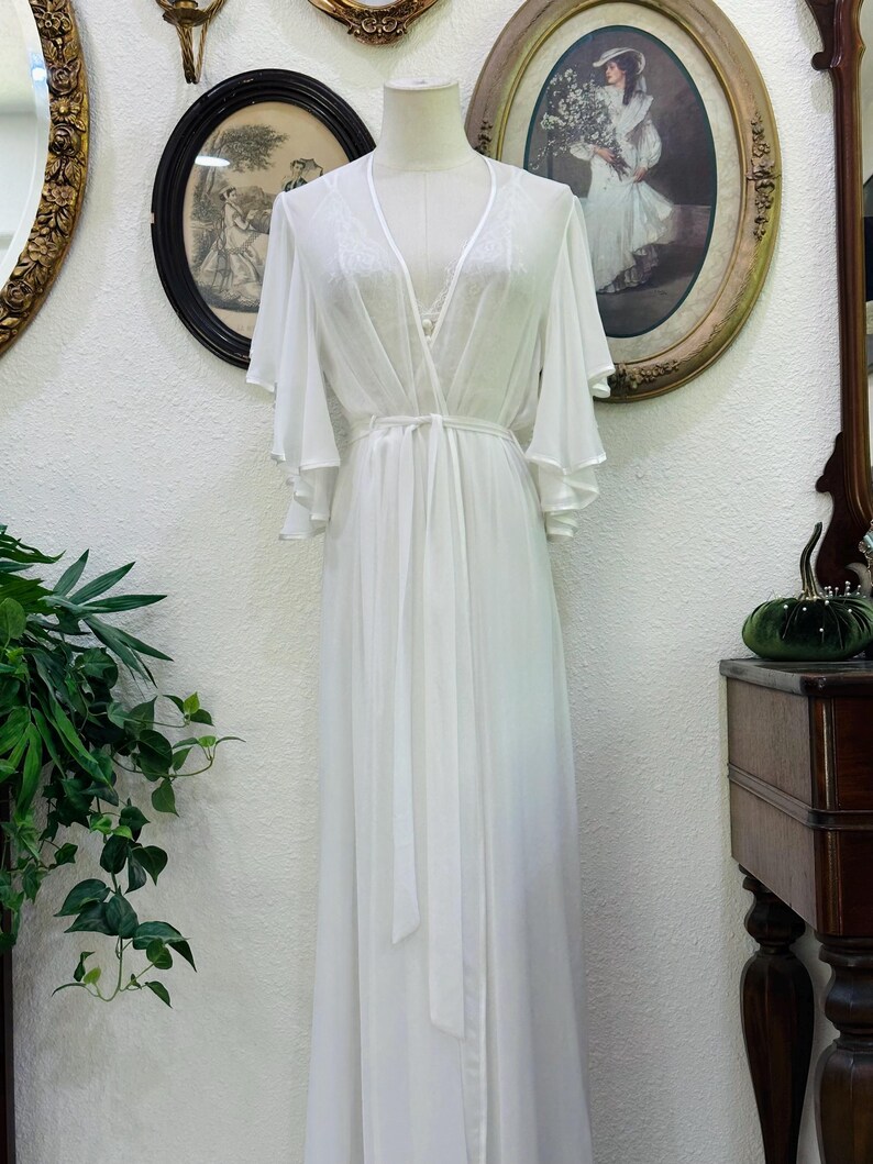 May include: A floor-length white robe featuring sheer fabric, a deep V-neckline, and wide, flowing bell sleeves. The garment includes a matching tie belt at the waist and delicate lace detailing along the neckline and shoulders, displayed on a mannequin.