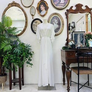 May include: A floor-length white robe featuring a wrap-style front, a tie waist, and wide, flowing flutter sleeves. The garment is displayed on a mannequin and is crafted from a sheer, lightweight fabric that drapes down to the floor.