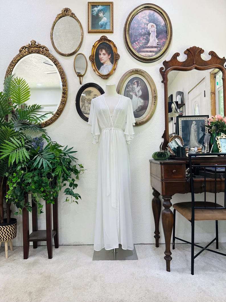 May include: A floor-length white robe featuring a wrap-style front, a tie waist, and wide, flowing flutter sleeves. The garment is displayed on a mannequin and is crafted from a sheer, lightweight fabric that drapes down to the floor.