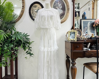 May include: A white lace dress on a mannequin. The dress has a round neckline, short sleeves, and a tiered skirt. The dress is displayed in a room with antique mirrors, framed portraits, and a wooden desk.