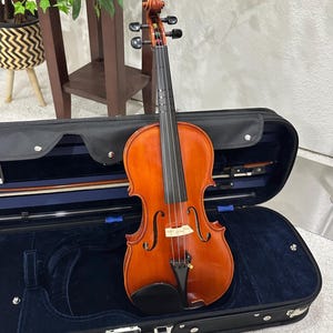 May include: A wooden violin with a warm, reddish-brown finish, resting in an open black case with a blue interior. The violin's strings and bow are visible, suggesting a musical instrument ready for play.