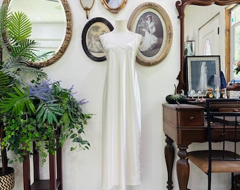 May include: A floor-length, cream-colored satin nightgown featuring thin spaghetti straps, a delicate lace trim along the V-neckline, and a subtle lace detail on the side of the skirt, displayed on a mannequin stand.