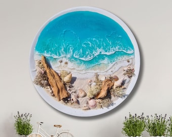 Könnte beinhalten: Ein rundes Harz-Kunstwerk, das eine Strandszene mit blauem Wasser, weißem Sand und Muscheln darstellt. Das Stück ist mit einem weißen Rahmen versehen.