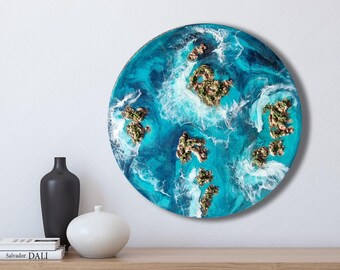 Könnte beinhalten: Runde Wandkunst mit einer lebendigen Ozeanszene in Türkis und Weiß. Mehrere goldbraune Inseln sind über dem Wasser verstreut. Das Kunstwerk wird an einer weißen Wand über einem Holzregal ausgestellt.