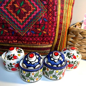 May include: Four small, hand-painted ceramic jars with lids. Two jars have blue lids with white floral designs, and two have white lids with red fruit designs. The jars are decorated with green leaves and yellow dots.