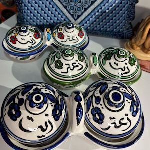 Può includere: Set di tre ciotole per doppio tuffo in ceramica con coperchi. Le ciotole sono decorate con intricati motivi floreali e calligrafia araba in blu, verde e rosso. I coperchi hanno un piccolo pomo in cima.