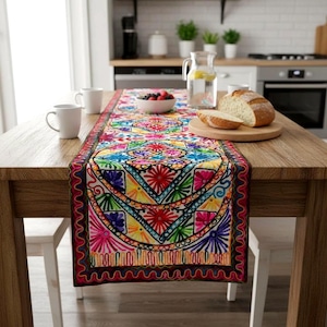 May include: A rectangular table runner featuring intricate, multi-coloured embroidery with geometric patterns in shades of red, blue, yellow, green, and purple. The runner is centred on a wooden table, accompanied by two white mugs and a loaf of bread.