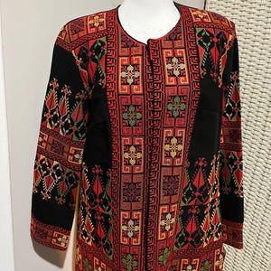 Peut inclure: Veste noire avec broderies complexes rouges, vertes et dorées. La veste présente un motif géométrique avec des motifs floraux et en losange. La veste à manches longues est présentée sur un mannequin, mettant en valeur son design et son savoir-faire.