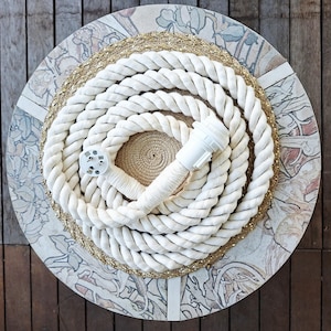 May include: A coiled white rope with a white electrical plug, on a round decorative surface. The surface has a floral pattern and a gold border. The rope is thick and appears to be made of natural fibres.