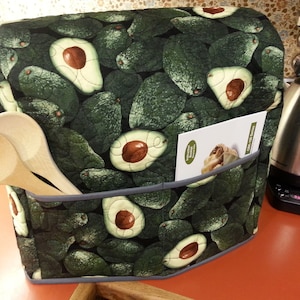 May include: A green and gray quilted kitchen appliance cover with a pattern of avocados. The cover has a pocket on the side that holds a recipe card. Two wooden spoons are in the pocket.