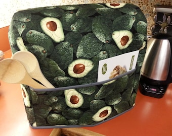 May include: A green and gray quilted kitchen appliance cover with a pattern of avocados. The cover has a pocket on the side that holds a recipe card. Two wooden spoons are in the pocket.