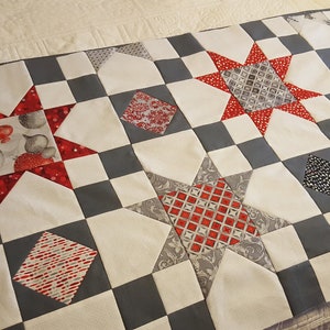 Pode incluir: Um edredom branco e cinza com um padrão de estrelas vermelhas e brancas. O edredom é feito de quadrados e diamantes. As estrelas estão no centro de cada quadrado. Os diamantes estão nos cantos de cada quadrado.