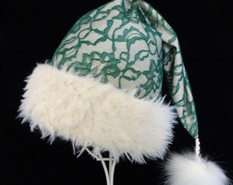 May include: A festive Santa hat with a green lace overlay and cream-colored faux fur trim. The hat features a fluffy white pom-pom at the tip and a matching fur band. The hat is displayed on a white stand against a black background.