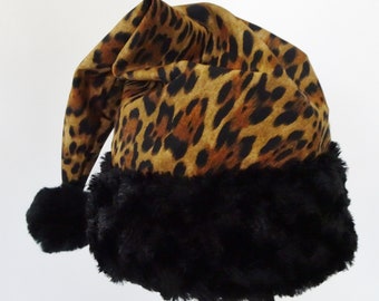May include: A Santa hat with a leopard print pattern in shades of brown and black. The hat has a black faux fur trim and two black pom-poms. The hat is made of fabric and is designed to be worn during the holiday season.