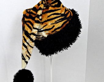 May include: A floppy hat featuring a vibrant orange and black tiger stripe pattern. The base of the hat is trimmed with a thick, textured black faux fur band. A large, round black pom-pom hangs from the pointed tip of the long, slouchy fabric crown.