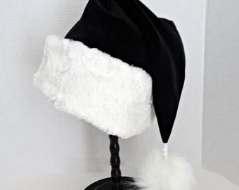 May include: A black Santa hat with a white faux fur trim and a matching pom-pom at the tip. The hat is displayed on a black stand against a white background. The hat's design is a classic holiday accessory.