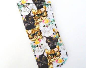 Op de afbeelding: Een stoffen brillenkoker met een herhalend patroon van witte, zwarte en oranje katten. Verschillende katten dragen een bril met een zwart montuur en kleine roze en gele bloemaccenten. De koker heeft een rechthoekige vorm met afgeronde hoeken.