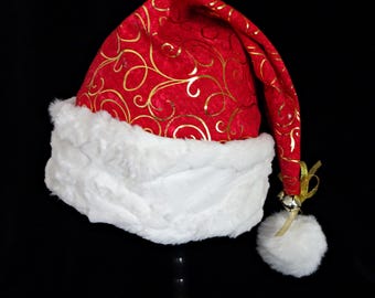 May include: A red Santa hat with gold swirl patterns and a white faux fur trim. The hat has a white pom-pom and a small silver bell attached with a gold ribbon. A festive Christmas accessory.