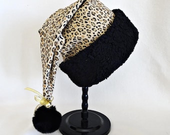 May include: A conical hat featuring a tan and black leopard print pattern with a wide, textured black cuff. The tip of the hat is adorned with a gold ribbon bow, a small metallic bell, and a fluffy black pom-pom, all displayed on a black stand.