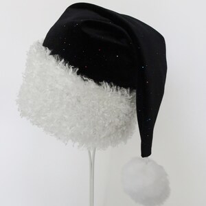 May include: A black Santa hat with a white, fluffy trim and a matching pom-pom. The hat is sprinkled with colorful glitter, adding a festive touch. The hat is displayed on a white stand against a white background.