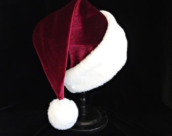 Può includere: Un cappello da Babbo Natale in velluto bordeaux intenso con un bordo spesso in pelliccia sintetica bianca soffice alla base e un pon-pon bianco abbinato fissato sulla punta. Il cappello è esposto su un supporto nero, mostrando la sua consistenza.