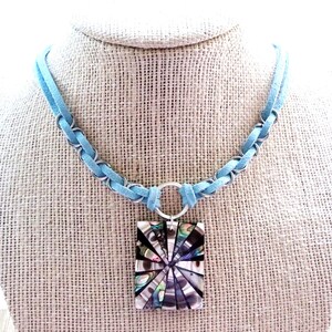 May include: A light blue suede cord necklace featuring silver-toned metal rings woven through the cord. A rectangular pendant hangs from a central silver ring, displaying a radial mosaic pattern of iridescent abalone shell segments.